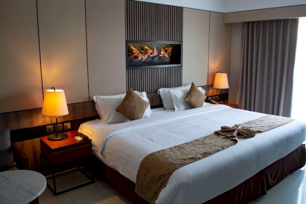 A cozy hotel room with a large bed, bedside tables with lamps, a wall-mounted fireplace, and a window with curtains.