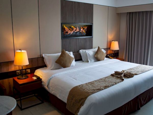 A cozy hotel room with a large bed, bedside tables with lamps, a wall-mounted fireplace, and a window with curtains.