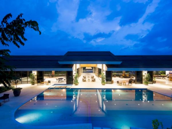 A luxurious building with a large swimming pool illuminated at night, lounge chairs around, and a partly covered area with seating.
