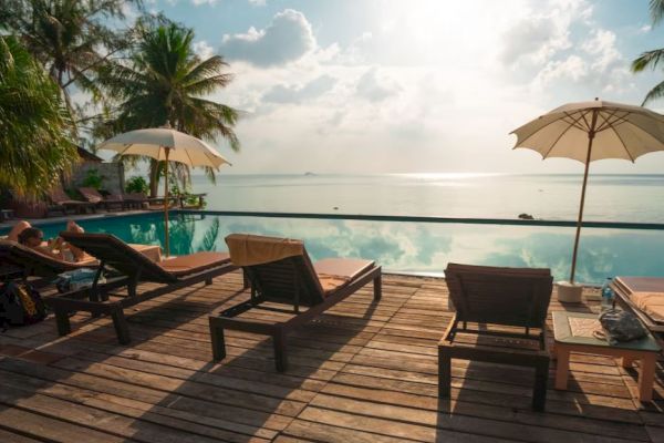 A serene poolside scene with lounge chairs, umbrellas, palm trees, and a view of the sea under a bright sky.