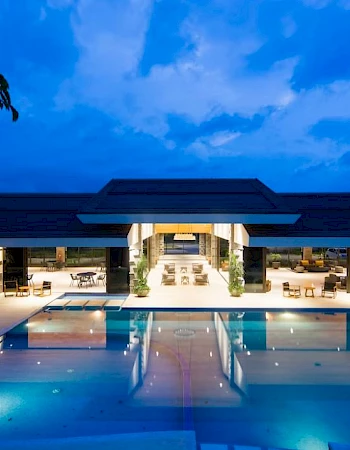 A modern, luxurious building with a large swimming pool, surrounded by lounge chairs and tables, set against a vibrant evening sky.