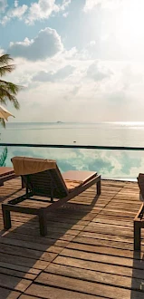 A serene poolside scene with lounge chairs, umbrellas, a wooden deck, and palm trees under a bright sky, overlooking a calm ocean view.