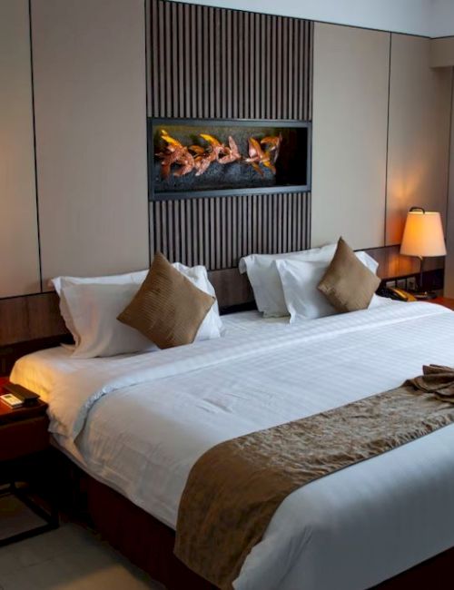 A well-lit hotel room with a large bed, two bedside tables with lamps, and a wall-mounted TV. The bed has beige pillows and a decorative towel.