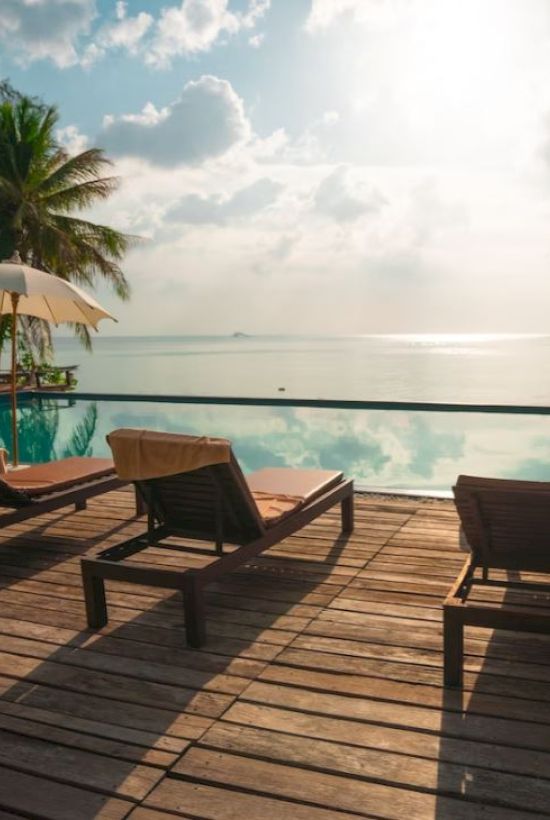 The image shows a serene poolside setting with lounge chairs, umbrellas, and palm trees, overlooking a calm body of water under a partly cloudy sky.