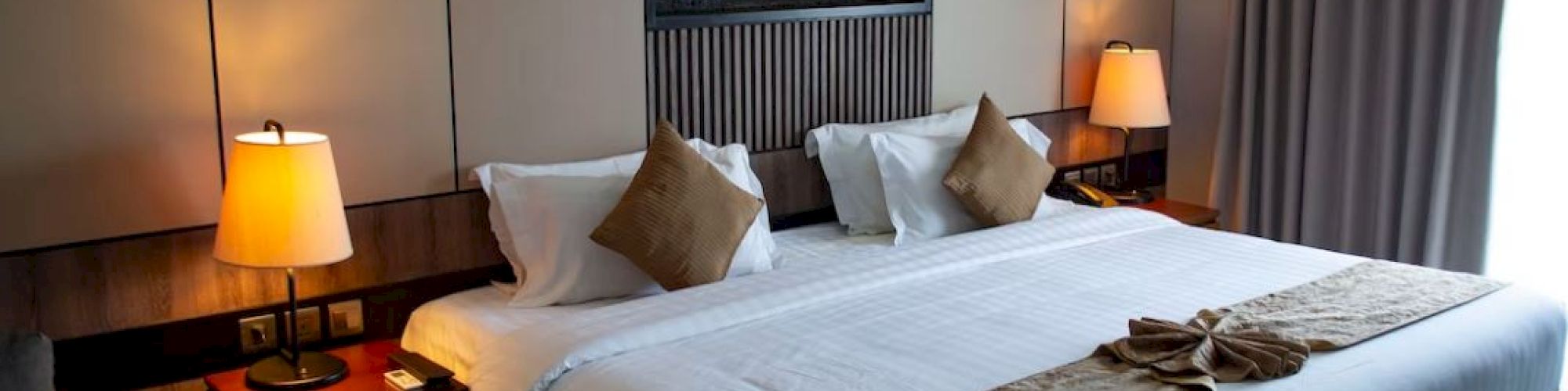 A well-lit hotel room features a large bed with white linens and brown accents, flanked by two bedside tables and lamps, with a fire display above.
