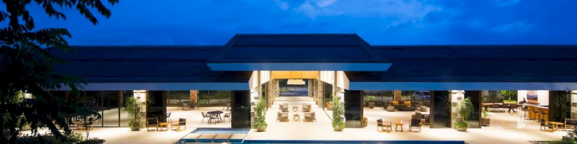 A luxurious indoor-outdoor space with large swimming pool, seating areas, and modern architectural design, illuminated beautifully at twilight.