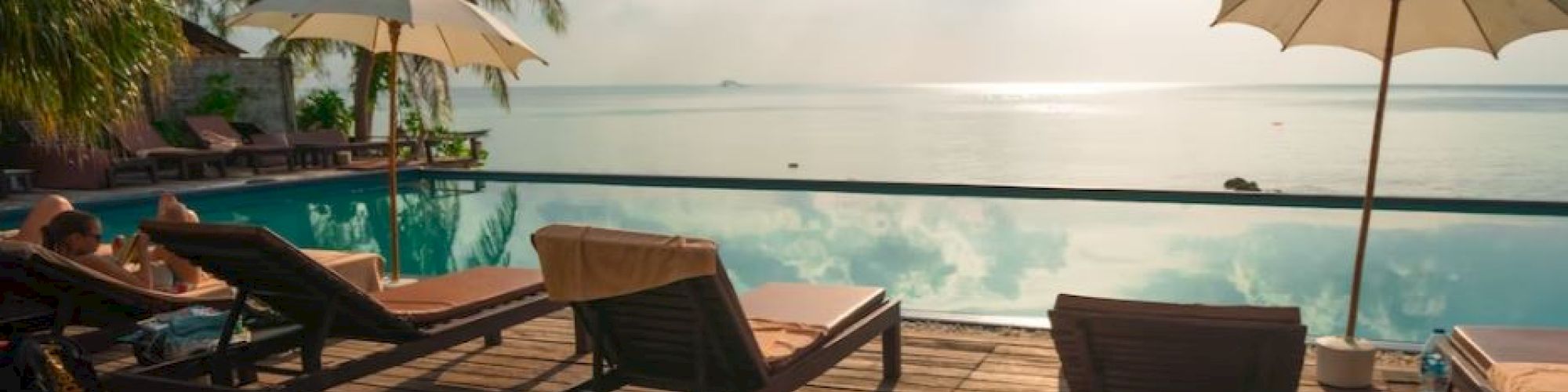 A serene poolside scene with lounge chairs, umbrellas, palm trees, and a stunning view of the ocean under a partly cloudy sky during sunset.