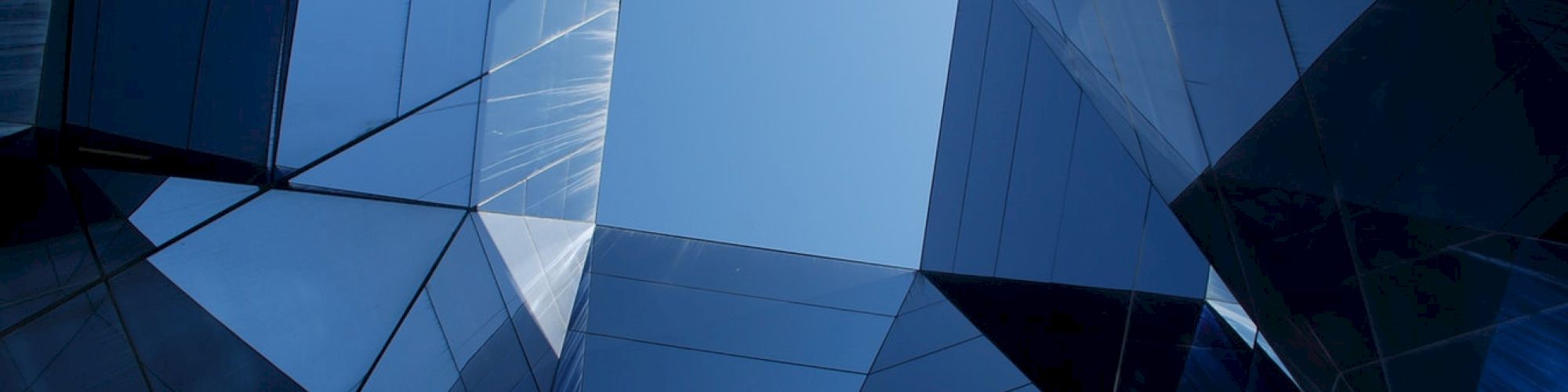 This image shows a view looking up through a geometric structure made of reflective glass panels, revealing a clear blue sky in the center of the frame.
