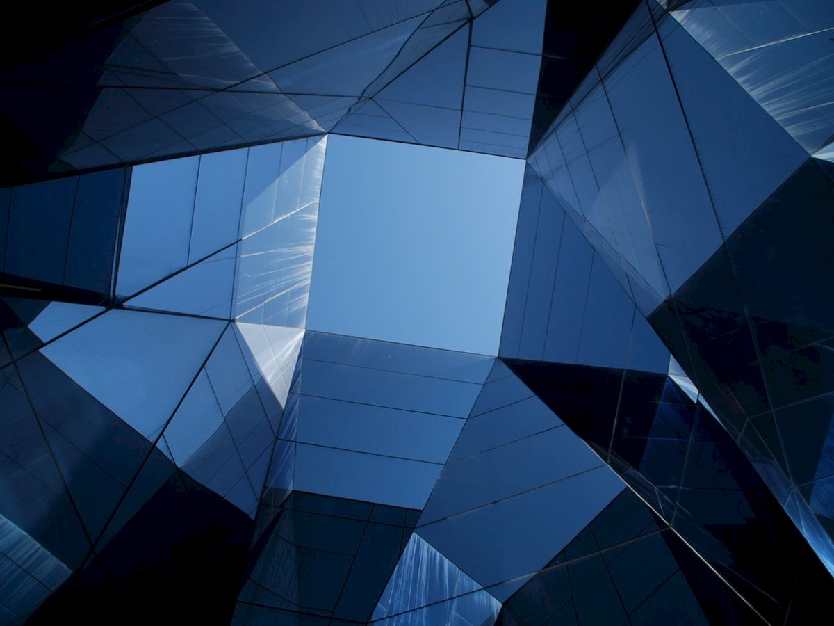 This image shows an abstract view of modern glass buildings meeting at an angular point, with reflections creating geometric patterns against a blue sky.