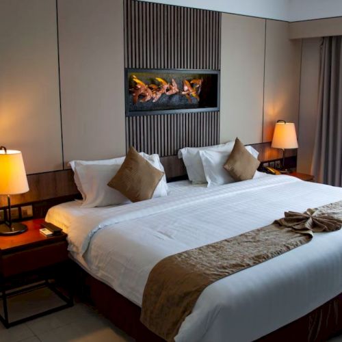 A neatly decorated hotel room features a large bed with white linens and brown accents, two bedside tables with lamps, and a wall-mounted fireplace.