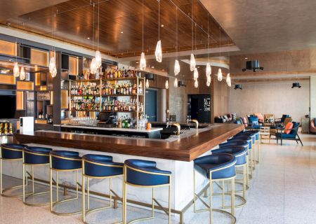 A modern bar with blue and gold stools, a well-stocked bar, unique hanging lights, and a seating area with couches in the background.