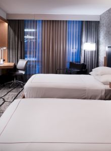 A modern hotel room features two neatly made beds, a flat-screen TV, a desk with a chair, and large windows with curtains.