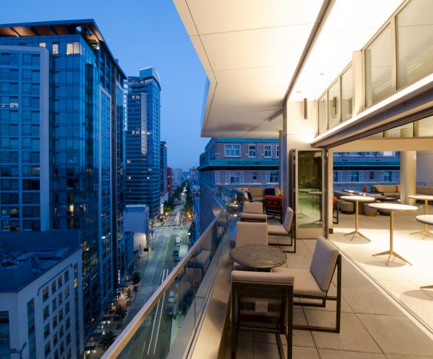 A modern cityscape at dusk with tall buildings and bright lights. A spacious balcony extends from a building with outdoor seating and tables.