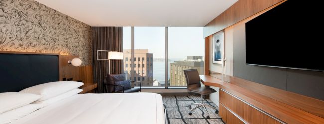 The image shows a modern hotel room featuring a large bed, a wall-mounted TV, a desk, a chair, and a window with a city view.