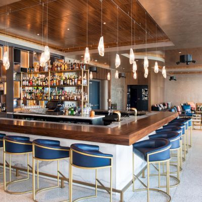 A modern bar with blue cushioned stools, a variety of liquors on shelves, stylish pendant lighting, and wooden accents throughout the space.