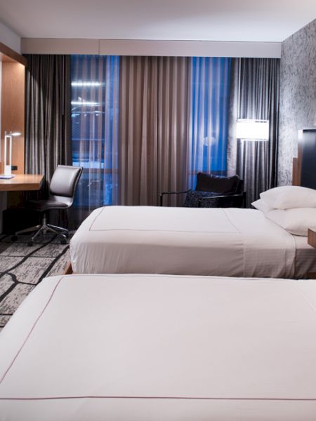 A modern hotel room with two beds, a TV on the wall, a desk by the window, and stylish decor, including a patterned wall and carpet.