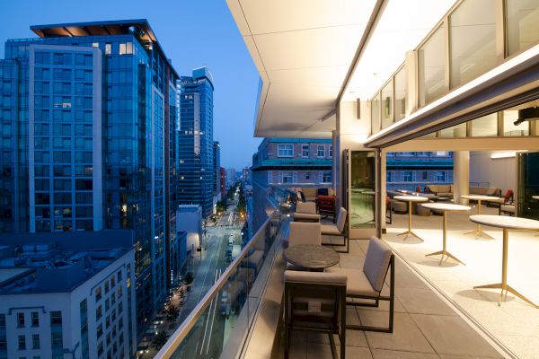 The image shows a modern outdoor balcony with seating and tables overlooking a cityscape of tall buildings at dusk.