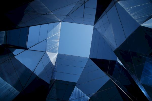 The image shows an upward view of an architectural structure with glass panels forming a geometric pattern, revealing the blue sky in the center.