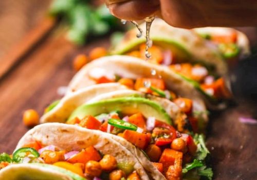 The image shows a hand squeezing lime juice over a row of chickpea and vegetable tacos, garnished with avocado slices, on a wooden surface.