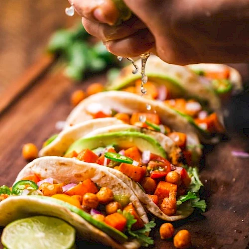 The image shows a hand squeezing lime juice over a row of chickpea and vegetable tacos, garnished with avocado slices, on a wooden surface.