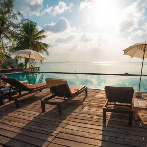 A serene beachfront scene with lounge chairs, umbrellas, a pool, palm trees, and an ocean view under a bright, partly cloudy sky ending the sentence.