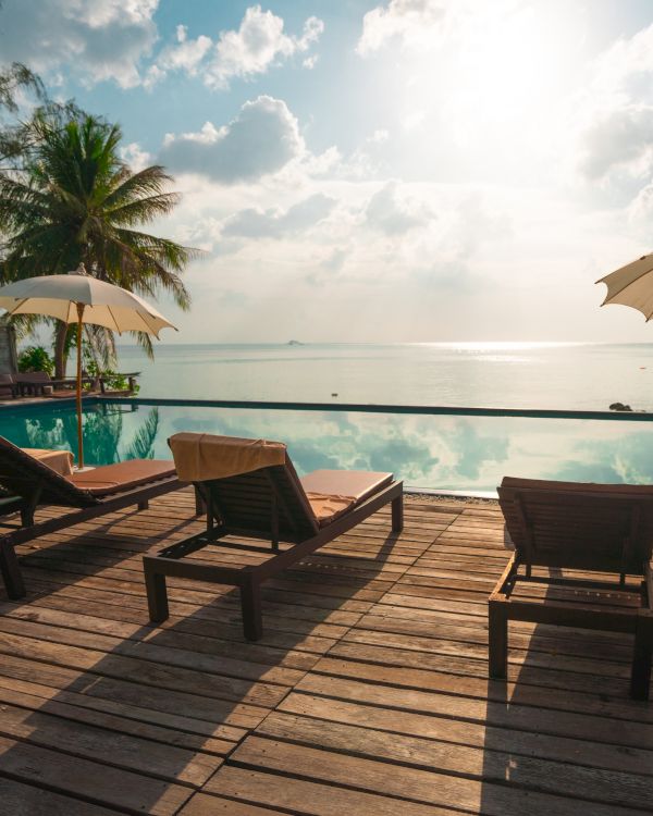 A serene beachfront scene with lounge chairs, umbrellas, a pool, palm trees, and an ocean view under a bright, partly cloudy sky ending the sentence.