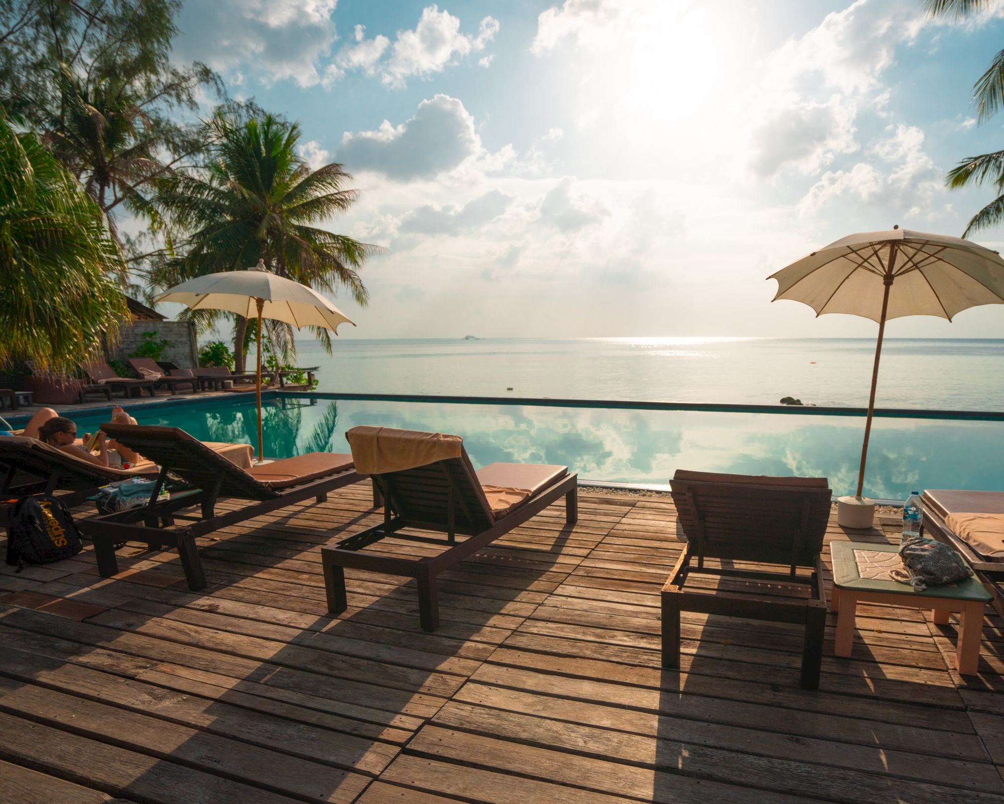 Sun loungers under umbrellas by an infinity pool, overlooking the ocean with a scenic view of the sun, sky, and palm trees in the background.