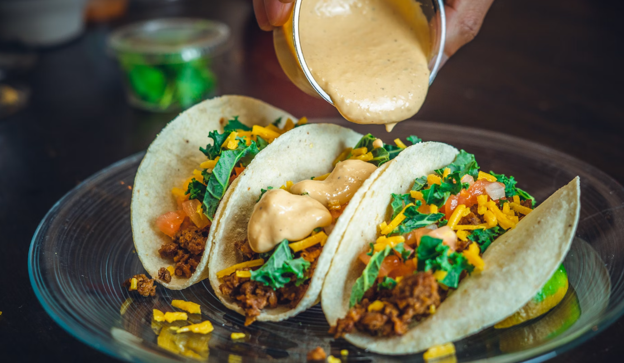 Three tacos are being topped with a creamy sauce, garnished with shredded cheese, greens, and diced tomatoes on a clear round plate.