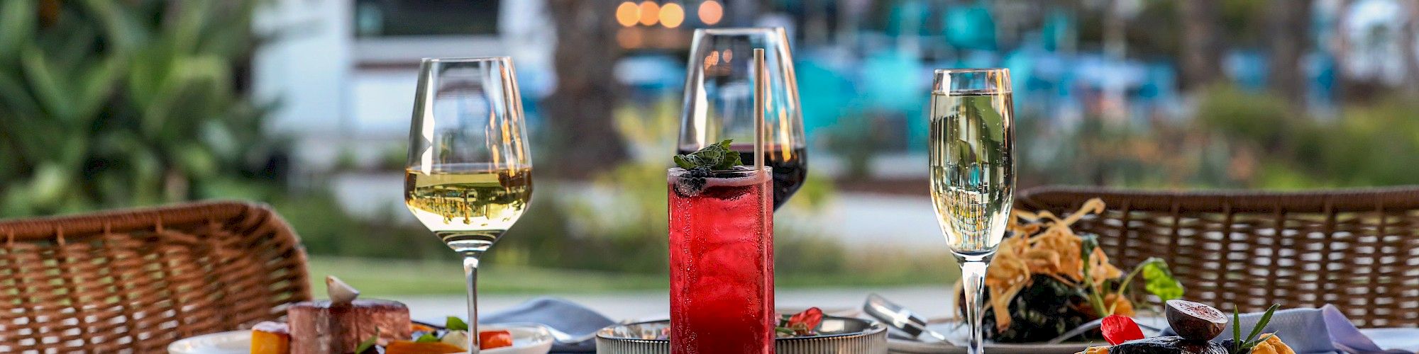 A beautifully set outdoor dining table features two plates of gourmet food, wine glasses, and a cocktail, with blurred lights in the background.