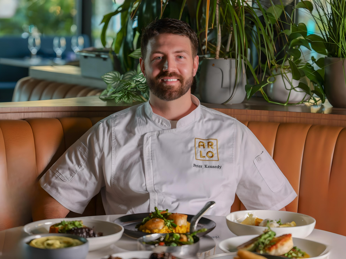 A chef sits at a restaurant booth with various plated dishes in front, wearing a uniform that reads "Arlo."