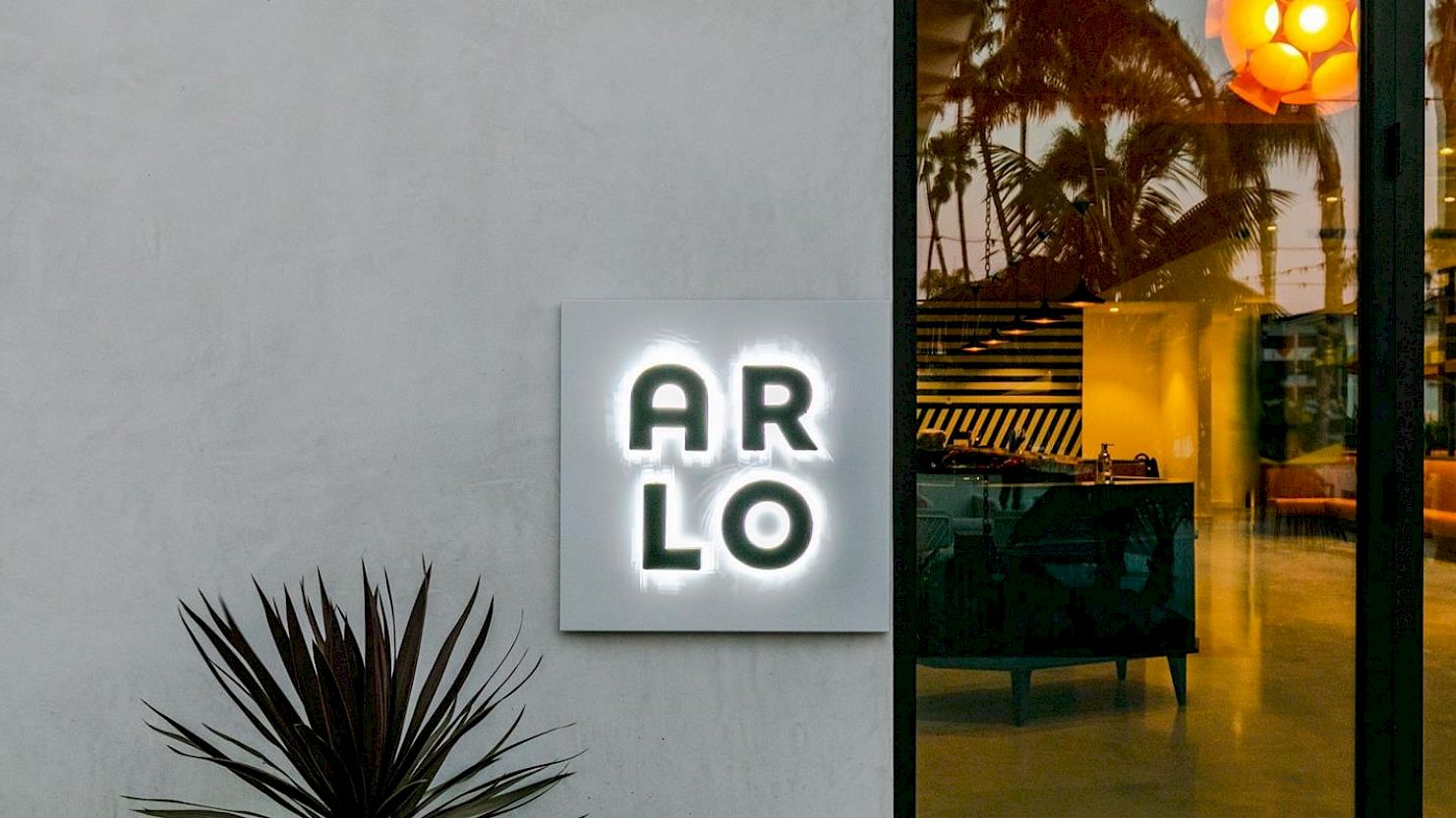 An illuminated sign reads "ARLO" on a building wall beside a glass door, with plants and reflections of palm trees visible in the background.
