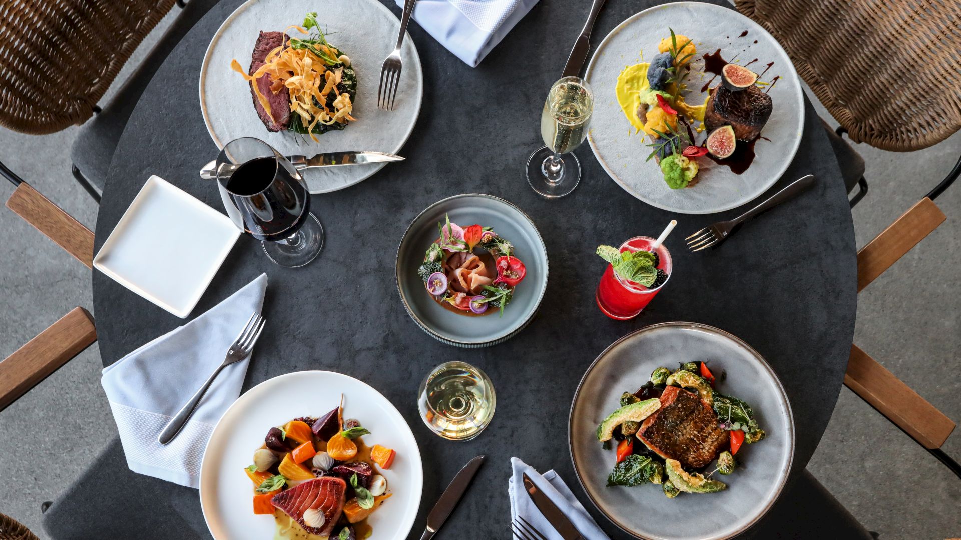 A table set with various gourmet dishes, wine, and cocktails, featuring elegantly plated meats, salads, and desserts, along with cutlery and napkins.