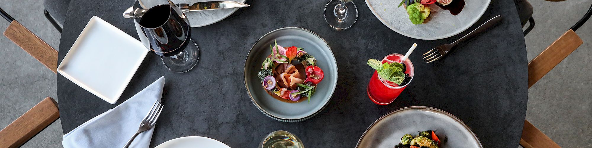 A table set with various gourmet dishes, wine, and cocktails, featuring elegantly plated meats, salads, and desserts, along with cutlery and napkins.