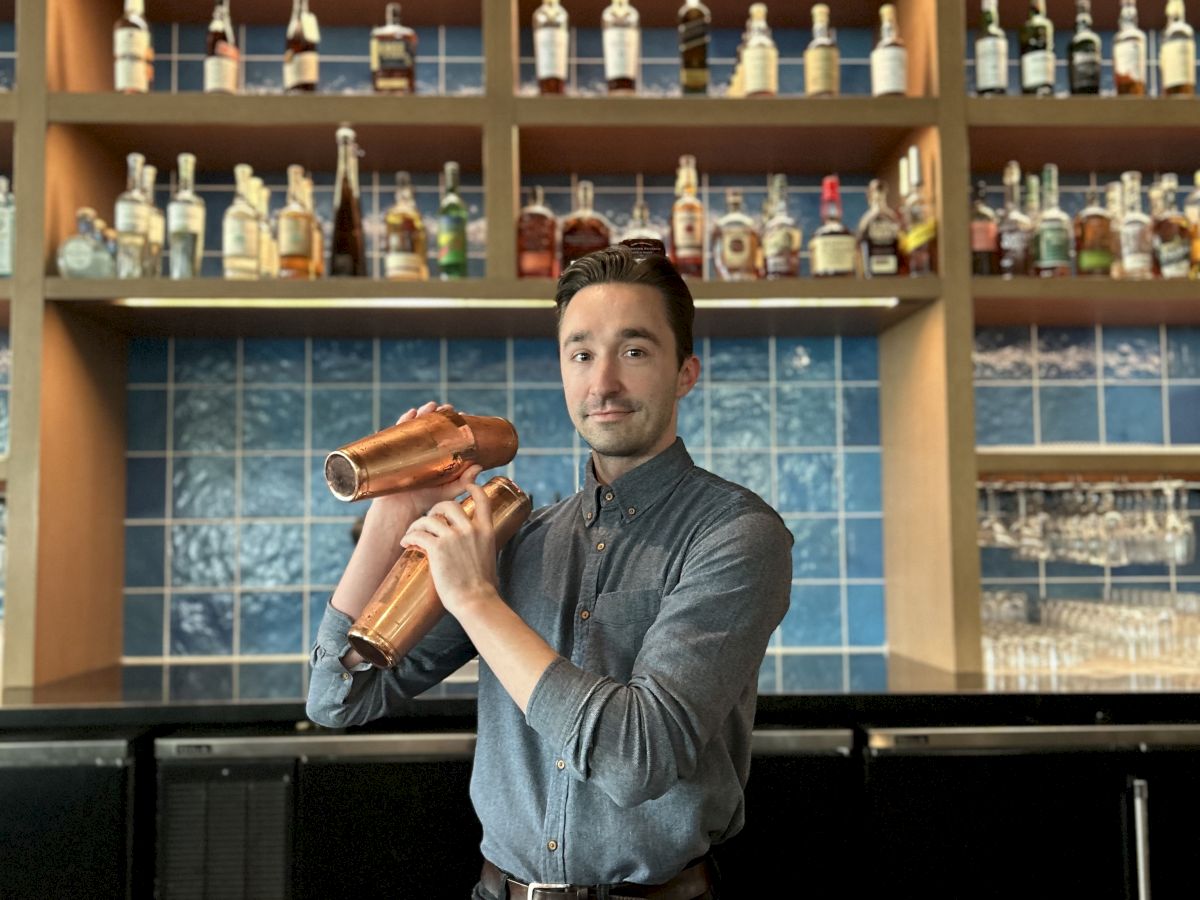 A bartender stands behind a bar shaking a cocktail shaker. In the background are shelves filled with various bottles of alcohol.