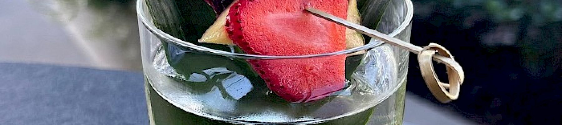 A clear glass filled with a cocktail garnished with pineapple leaves, a strawberry slice, and a piece of citrus and pineapple.