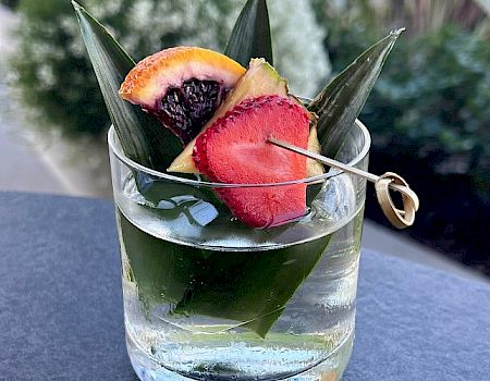 A clear glass filled with a cocktail garnished with pineapple leaves, a strawberry slice, and a piece of citrus and pineapple.