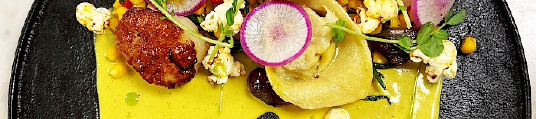 A gourmet dish on a black plate, featuring scallops, vegetables, microgreens, radish slices, and a bright yellow sauce drizzled creatively.