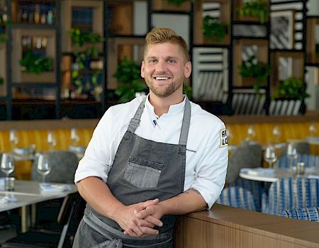 A smiling chef in a white uniform and gray apron stands in a modern restaurant with a stylish interior featuring plants and set tables.