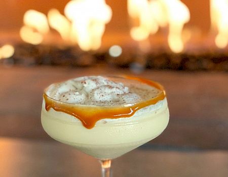 A creamy cocktail in a glass with caramel drizzling down the side, topped with whipped cream and sprinkled spices, set against a background of flames.