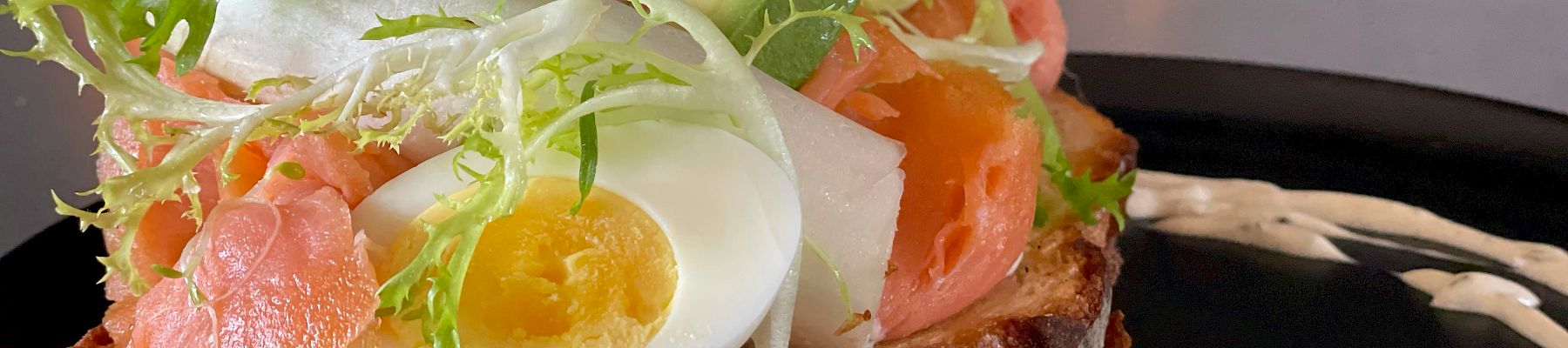 A toasted slice of bread topped with leafy greens, a boiled egg, tomato slices, and smoked salmon, garnished with a creamy dressing on a black plate.