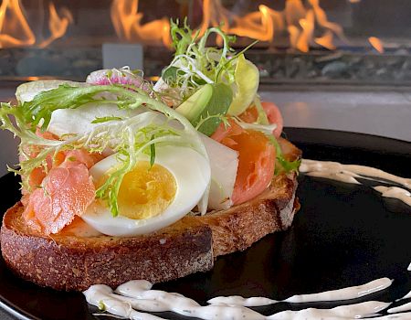 A toasted slice of bread topped with leafy greens, a boiled egg, tomato slices, and smoked salmon, garnished with a creamy dressing on a black plate.