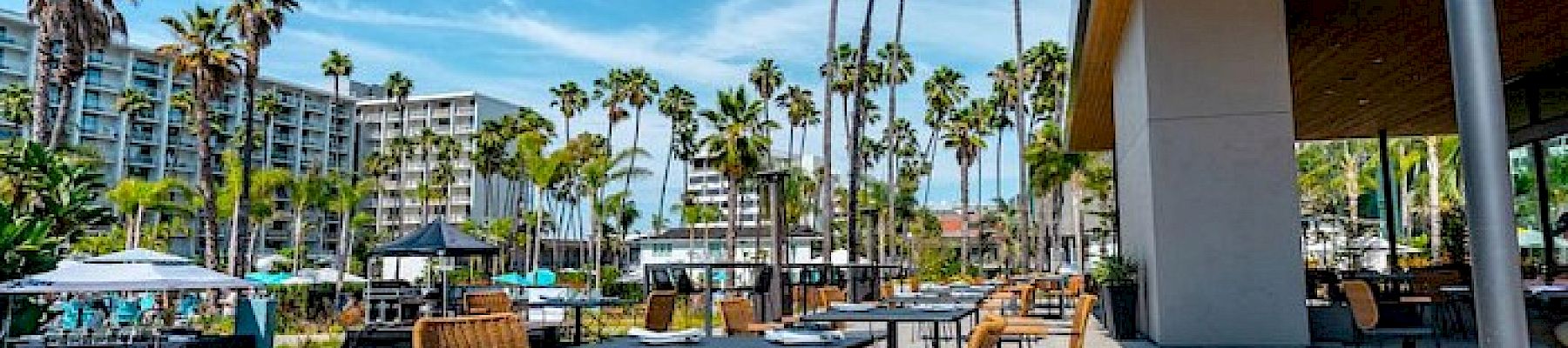 An outdoor patio area with tables and chairs, surrounded by tall palm trees and nearby buildings under a clear blue sky, creating a relaxing atmosphere.