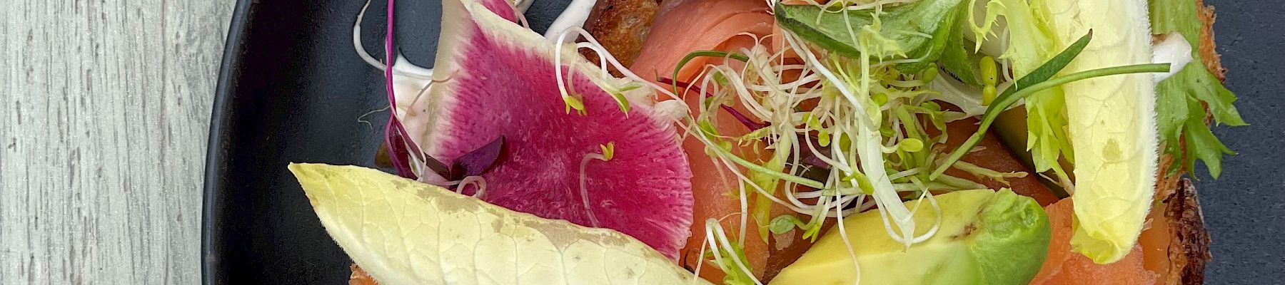 A black plate with a dish containing smoked salmon, avocado, boiled egg, radish, and greens, garnished with a white sauce.