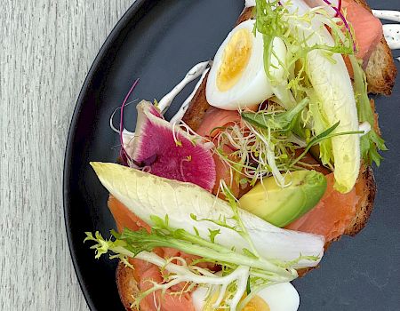 A black plate with a dish containing smoked salmon, avocado, boiled egg, radish, and greens, garnished with a white sauce.