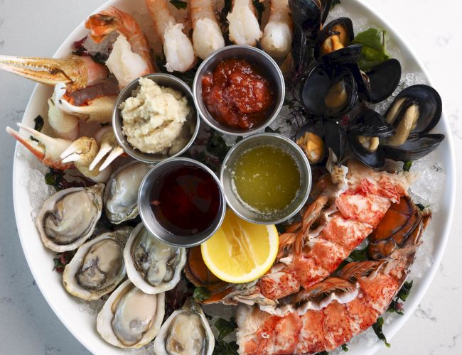 A seafood platter with shrimp, oysters, lobster, and mussels, accompanied by lemon wedges and various dipping sauces in small bowls on a white plate.