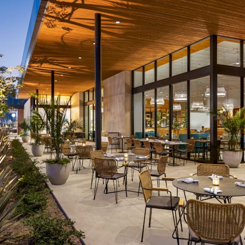 An outdoor restaurant with wicker chairs and tables, surrounded by plants, set under a wooden roof with glass walls leading inside.