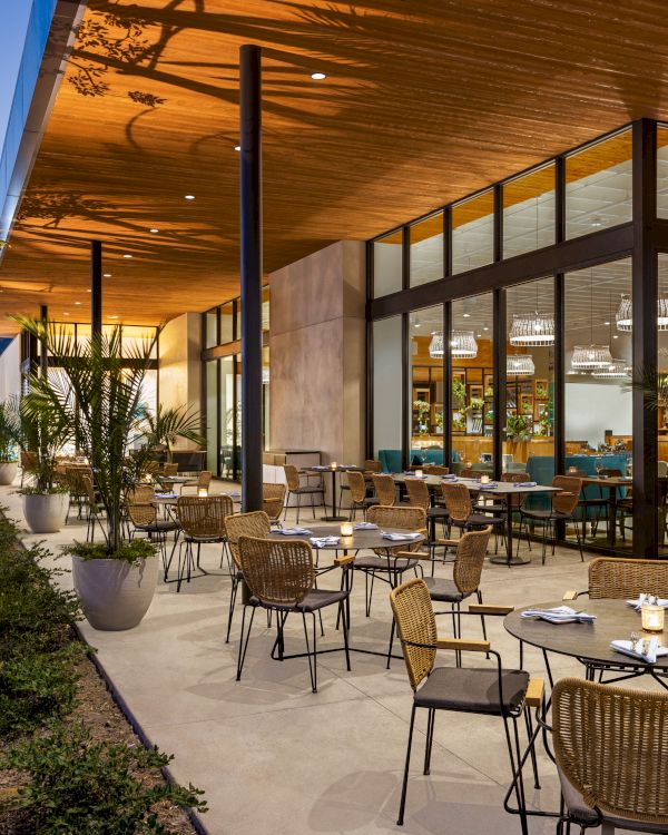 An outdoor restaurant with wicker chairs and tables, surrounded by plants, set under a wooden roof with glass walls leading inside.