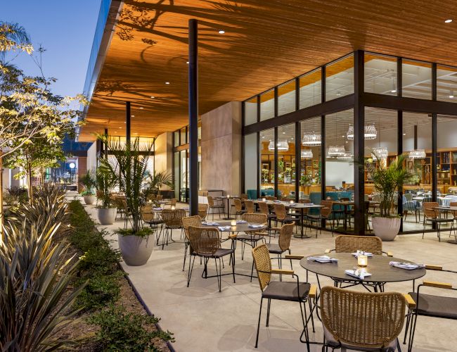 An outdoor restaurant with wicker chairs and tables, surrounded by plants, set under a wooden roof with glass walls leading inside.