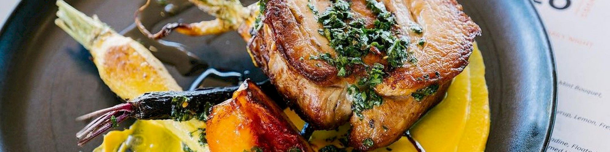 A beautifully plated dish featuring roasted meat, colorful vegetables, and a vibrant yellow sauce, served on a black plate with a menu beside it.