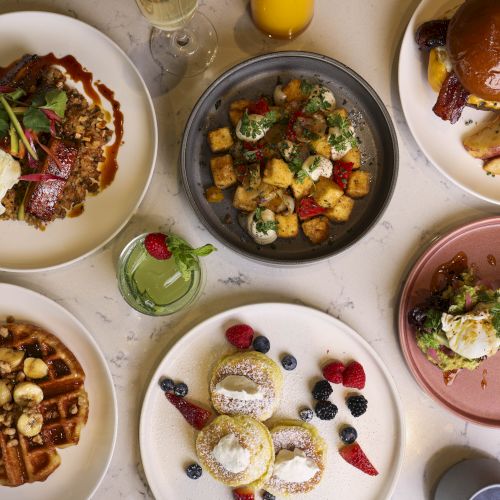 This image shows a spread of breakfast or brunch dishes, including waffles with bananas, stuffed pancakes, eggs benedict, roasted potatoes, and a sandwich.